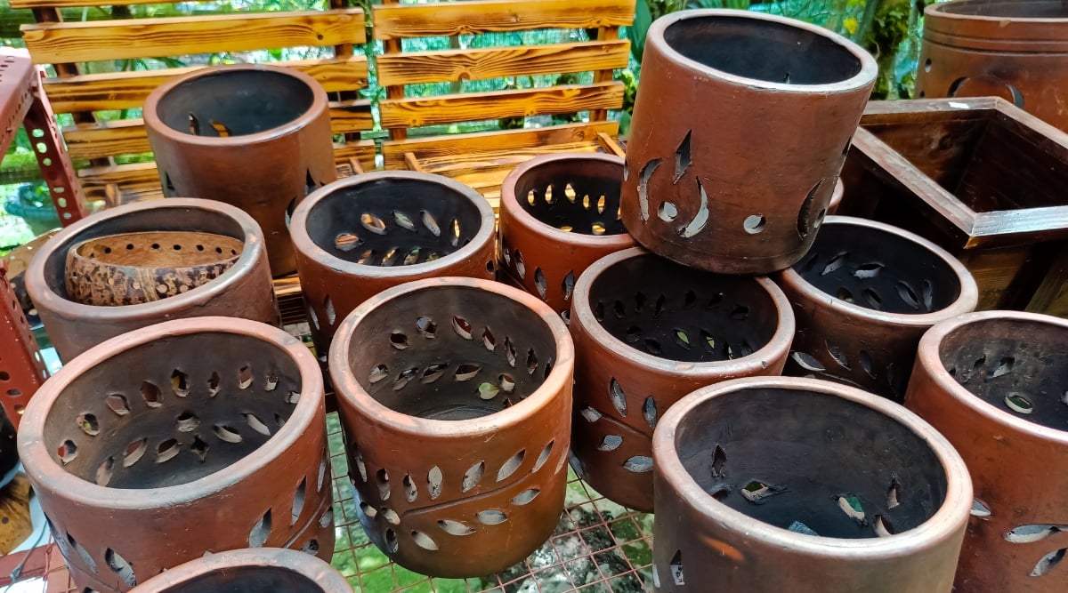 Neatly arranged clay pots designed for orchids are shown. They are adorned with decorative perforations, adding an artistic touch to their rustic charm. Placed against a garden screen backdrop, these pots harmoniously blend with the natural setting, creating a picturesque display.